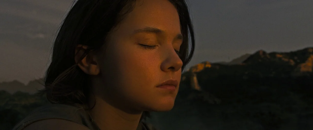 Alien: Romulus (2024) movie still: closeup — A person stands in front of a mountain range during the golden hour sunset. The subject has their eyes…
