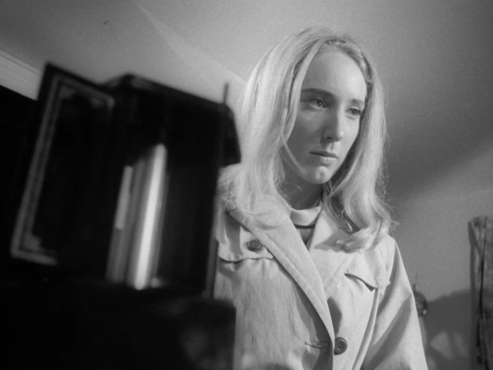 Night of the Living Dead (1968) movie still: medium — A woman stands in a dimly lit interior space with a blurred dark object nearby. The subject…