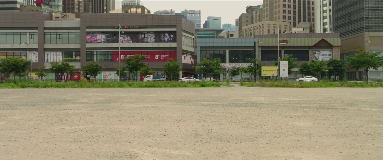 Psychokinesis (2018) movie still: establishing — A vast, unpaved gravel lot sits in front of a modern commercial city block. Multi-story buildings…