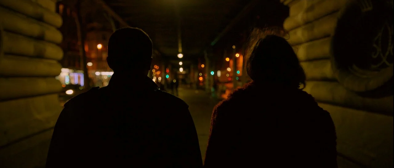 Love (2015) movie still: medium — Two figures walk down a dark stone bridge at night under distant glowing lights. The scene shows the backs of the…