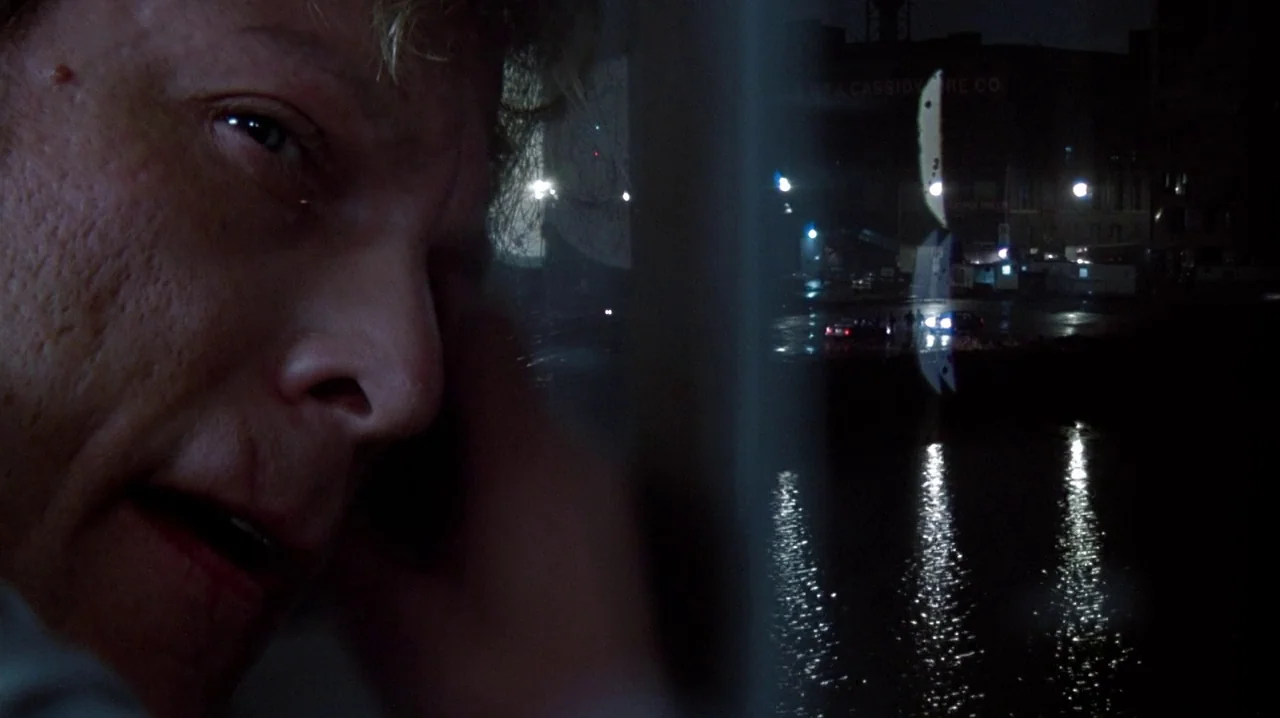 Thief (1981) movie still: closeup — A man's face is visible in close proximity to a window at night. Beyond the glass, a dark industrial harbor area…