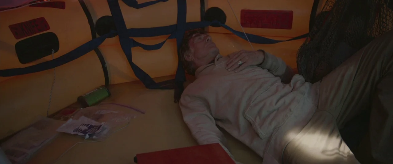 All Is Lost (2013) movie still: medium — An older man rests inside the cramped interior of an orange inflatable life raft. Supplies and netting are…