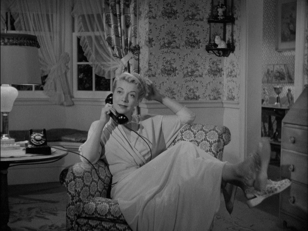 D.O.A. (1949) movie still: medium — A 1950s interior with floral wallpaper and patterned furniture. A blonde woman in a robe relaxes in an armchair…