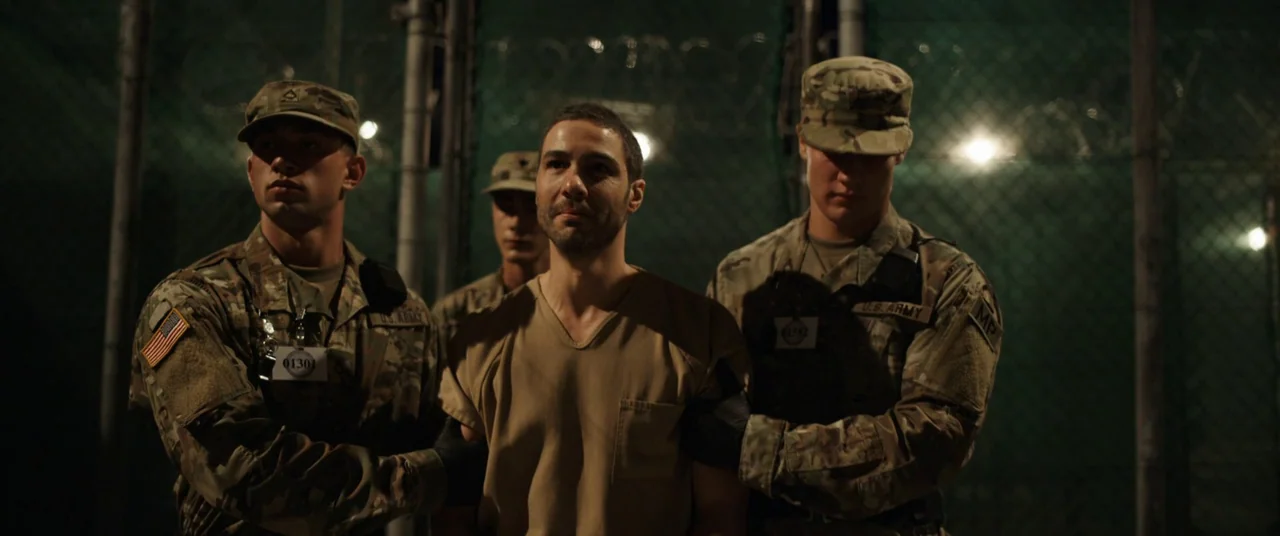 The Mauritanian (2021) movie still: medium — A dark outdoor military detention area is surrounded by high chain-link fences with barbed wire. Two…