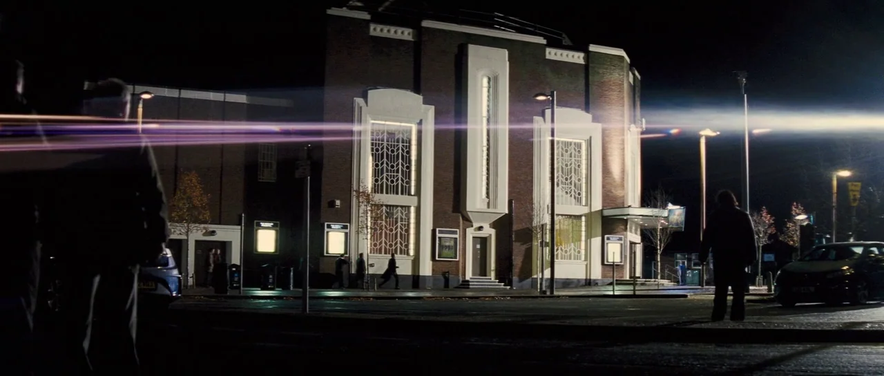 The World’s End (2013) movie still: wide — A brick art deco building stands illuminated by streetlights under a dark night sky. Several figures walk…