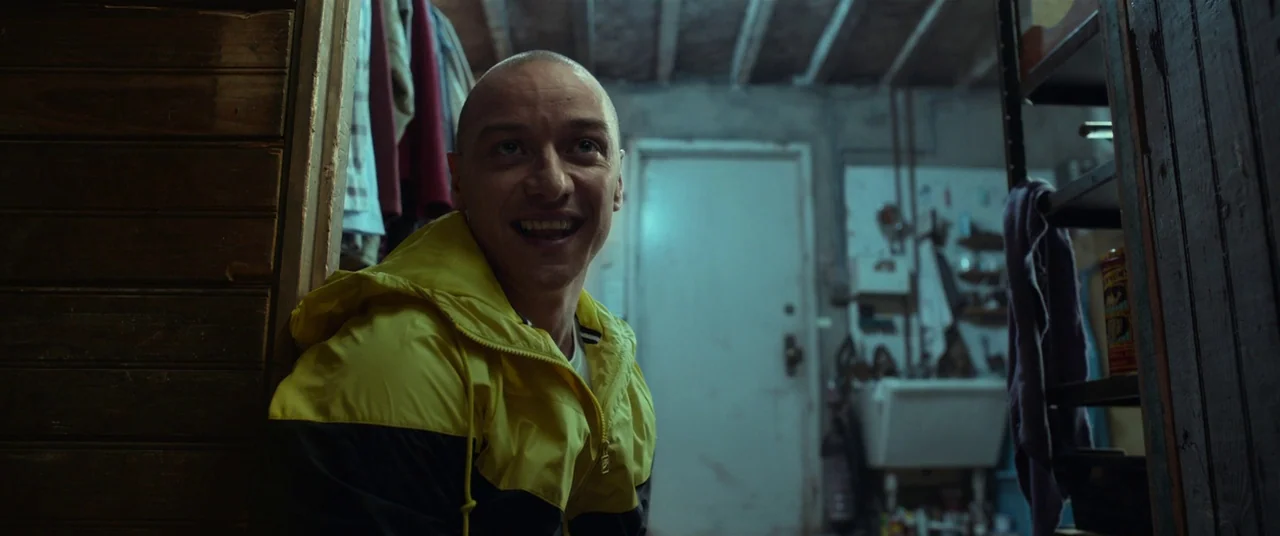 Split (2016) movie still: medium — A cluttered, dimly lit basement with a white utility sink and exposed ceiling joists. A man in a yellow and black…
