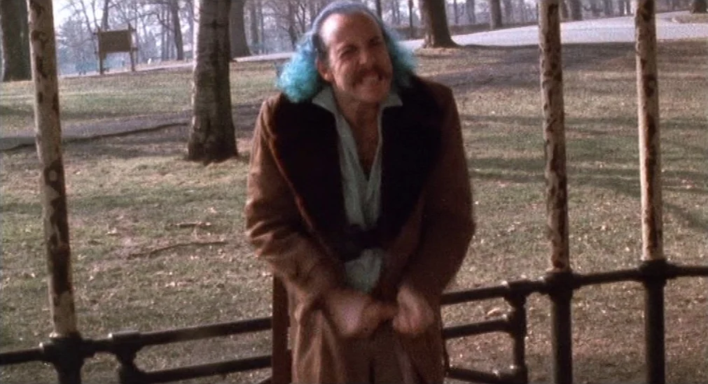 Pink Flamingos (1972) movie still: fullbody — A man stands inside a wooden gazebo structure located in a grassy park setting. The man clutches his…