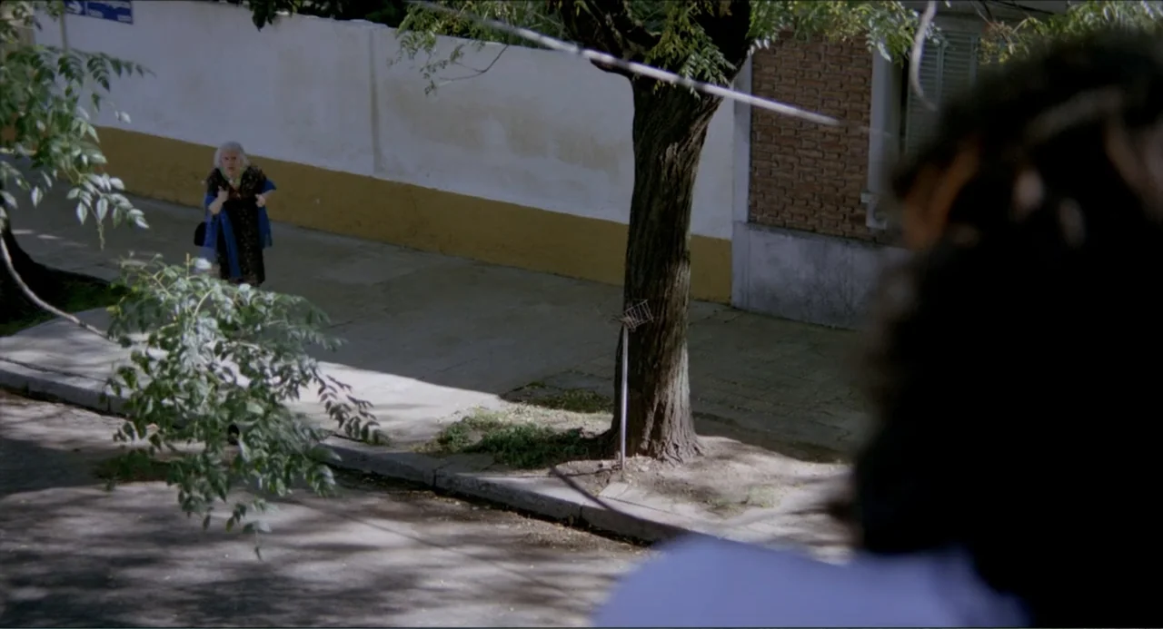 Waiting for the Hearse (1985) movie still: wide — A sunlit residential sidewalk features a long white wall and a single mature tree. An elderly…