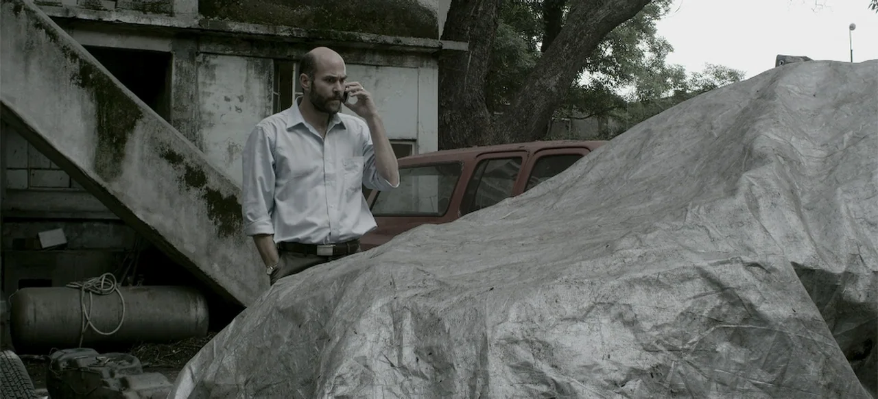 About the Deaths (2016) movie still: medium — A neglected outdoor area features a concrete structure and a car under a tarp. A bald man wearing a…