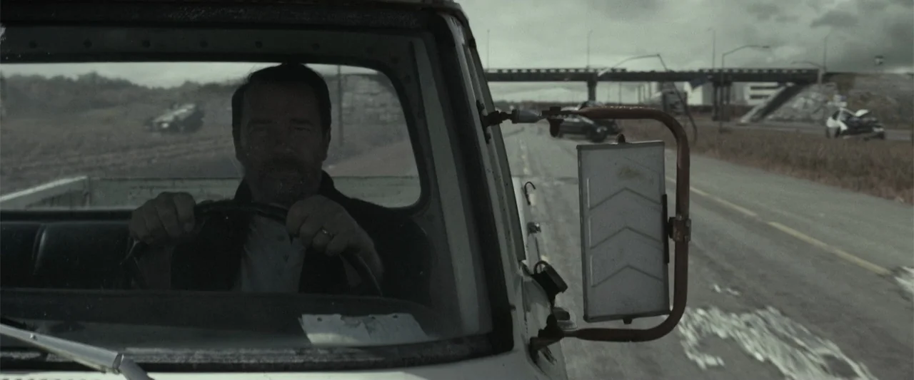 Maggie (2015) movie still: medium — A man drives a truck on a highway under an overcast sky. The vehicle interior shows a metallic side mirror and a…