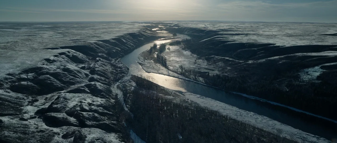 The Revenant (2015) movie still: establishing — A vast river winds through a snow-dusted valley flanked by dark, rugged hillsides. The low winter…