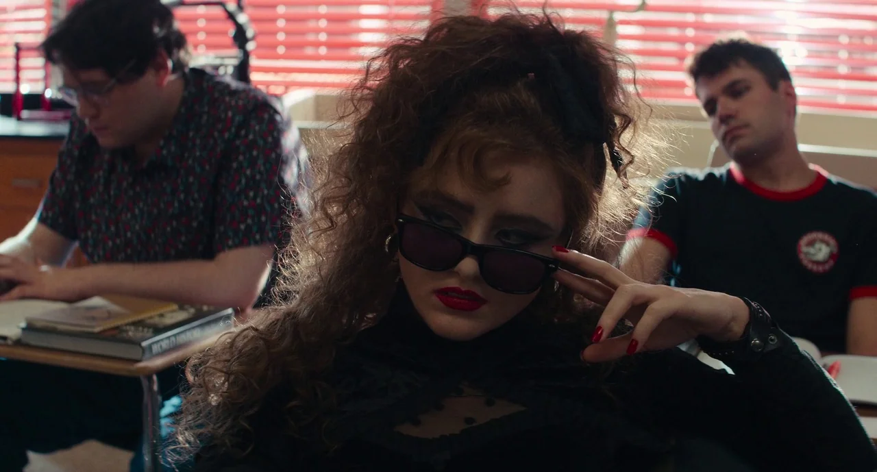 Lisa Frankenstein (2024) movie still: medium — A student sits in a classroom during the day with red window blinds visible behind. A teenage girl…