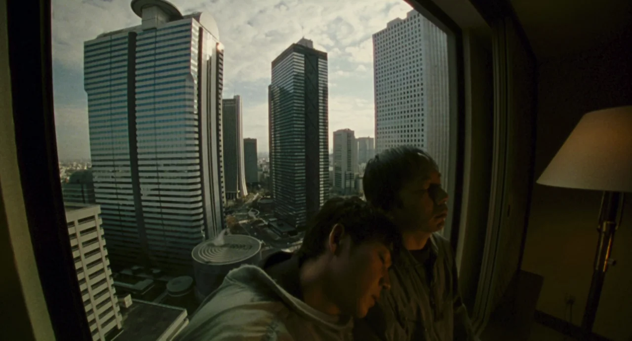 Ichi The Killer (2001) movie still: medium — A high-rise hotel room window overlooks a dense metropolitan district of large office towers. Two men…
