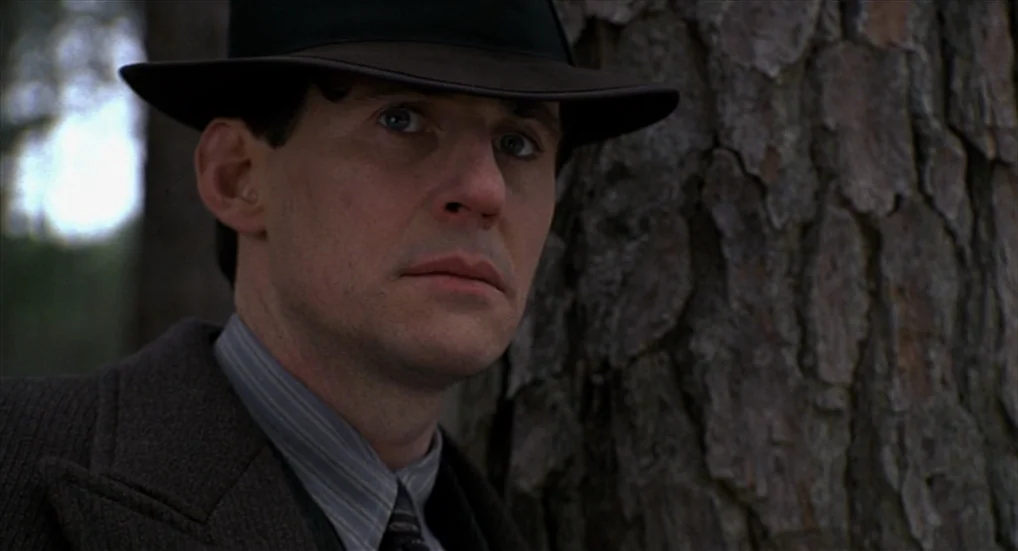 Miller’s Crossing (1990) movie still: closeup — A man stands next to a large pine tree in a wooded environment. He looks away from the camera into…