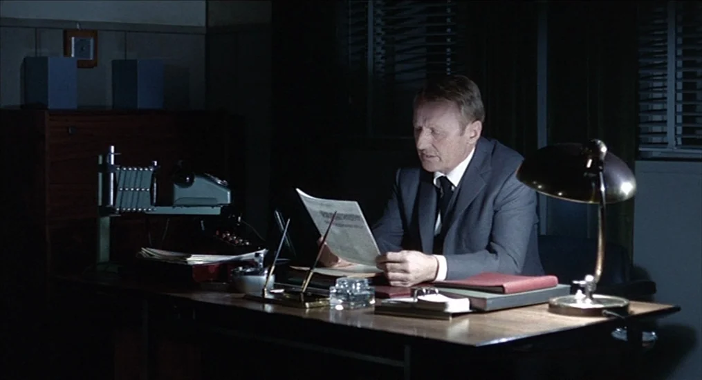 Le Cercle Rouge (1970) movie still: medium — A dimly lit room contains a large dark wood desk with various papers and equipment. A middle-aged man…