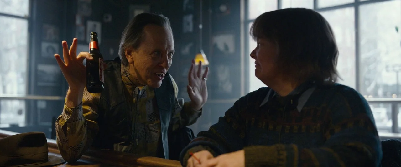 Can You Ever Forgive Me? (2018) movie still: medium — A dimly lit bar with framed photos on the dark wall behind a counter. An older man sits at the…