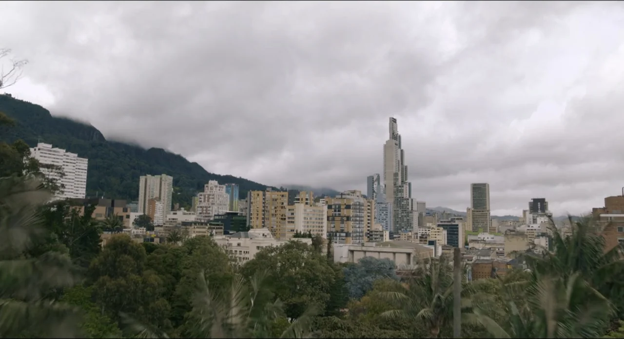 The Beauty and the Moody (2022) movie still: establishing — A dense urban skyline nestled against a large forested mountain under overcast skies.…