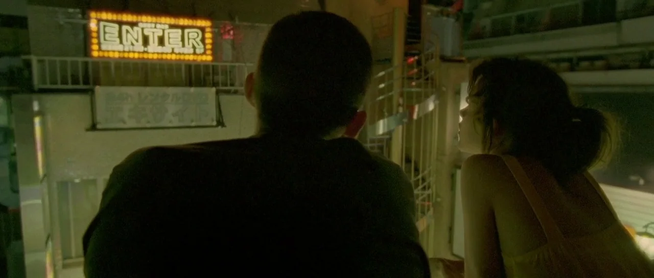 Enter The Void (2009) movie still: medium — A man and a woman stand on an exterior balcony at night looking across. A large glowing yellow 'ENTER'…