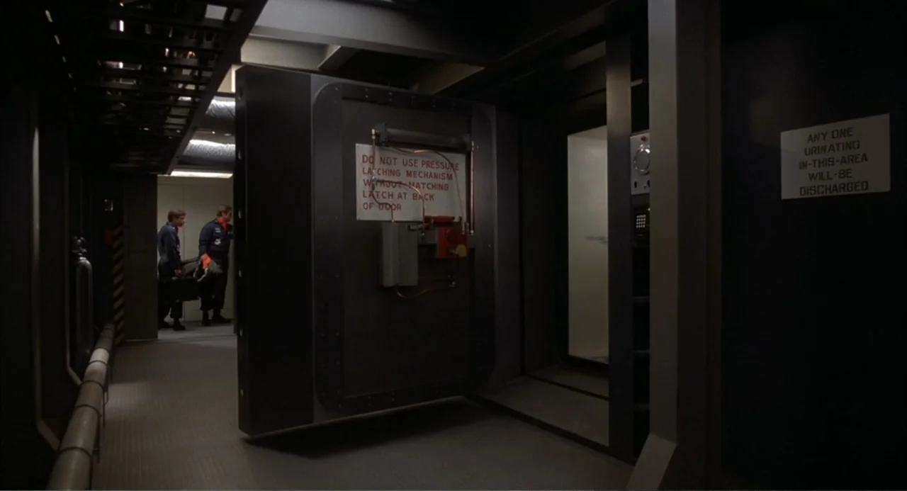 WarGames (1983) movie still: wide — A utilitarian industrial passageway features a large open steel vault door with warning signage. Two workers…