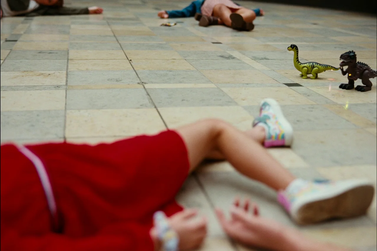 Bugonia (2025) movie still: medium — Three children lie spread out on a large-format light-colored stone floor. Two plastic dinosaur toys stand on…