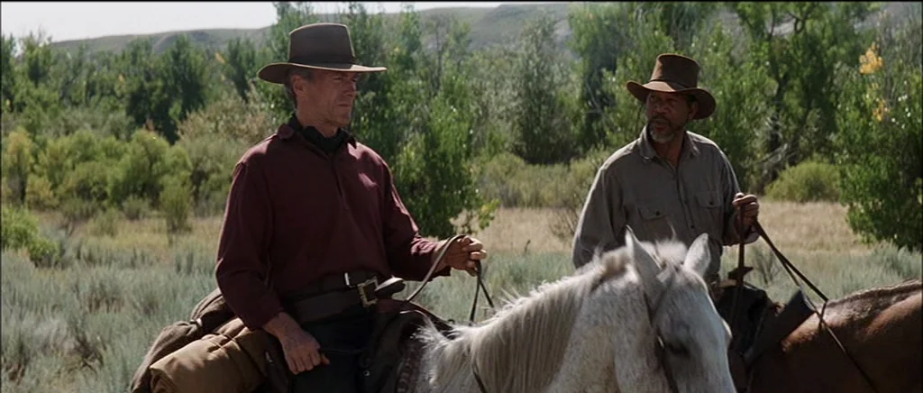 Unforgiven (1992) movie still: medium — Two men ride on horseback across a grassy expanse filled with green shrubs. The riders move together while…