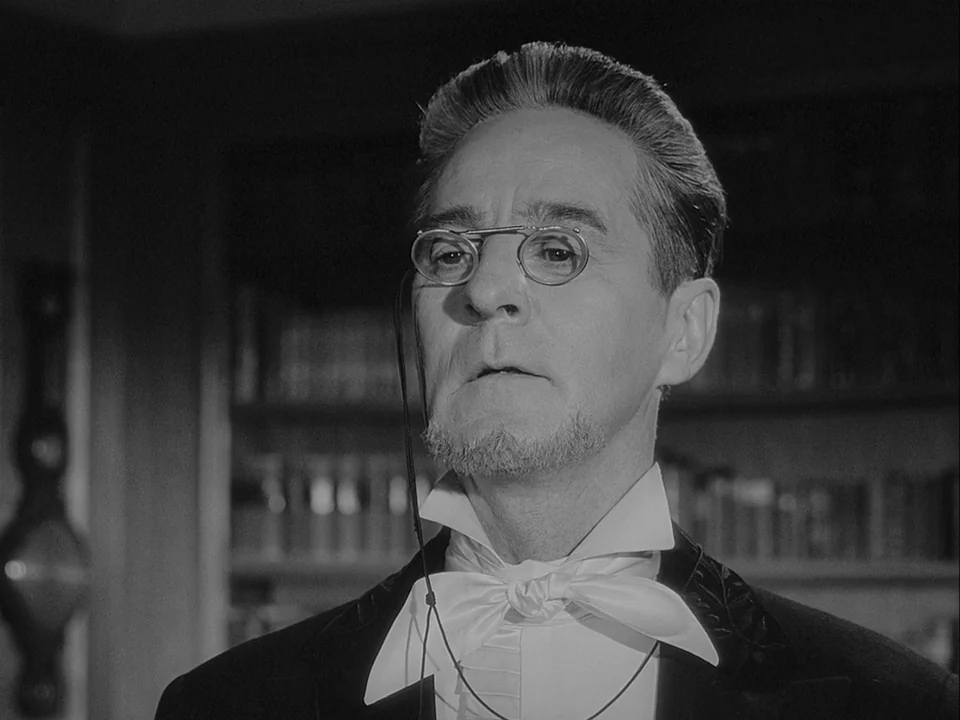 The Magician (1958) movie still: closeup — A dark paneled library contains rows of books on shelves visible in the background. A formally dressed…