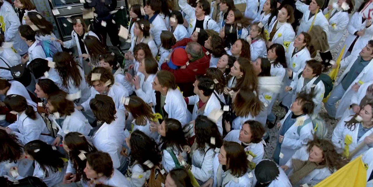 The Flower of My Secret (1995) movie still: wide — A dense crowd of students in white lab coats gathers in an outdoor plaza. Many individuals look…