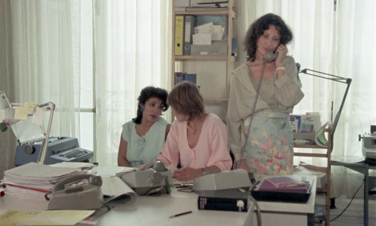 The Green Ray (1986) movie still: medium — An office environment with stacks of papers and clerical equipment on a white desk. Three women work…