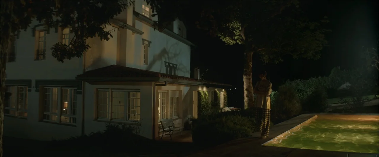 Tell Me Softly (2025) movie still: wide — A two-story house stands at night next to an illuminated swimming pool. A person in pajamas stands on the…