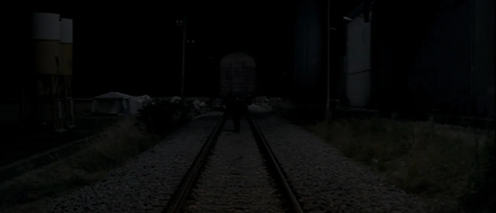 Time Of The Wolf (2003) movie still: wide — Train tracks at night with industrial structures visible in the distance. A single figure walks down the…