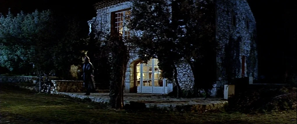Love Unto Death (1984) movie still: wide — A two-story stone house sits in darkness with trees partially obscuring its stone walls. A person stands…