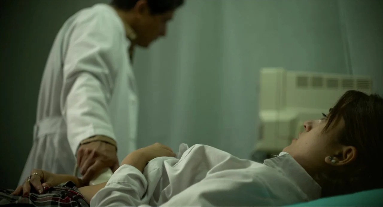 Heartbeat (2008) movie still: medium — An examination room with a patient lying on a bed while a doctor works. The scene centers on a medical…