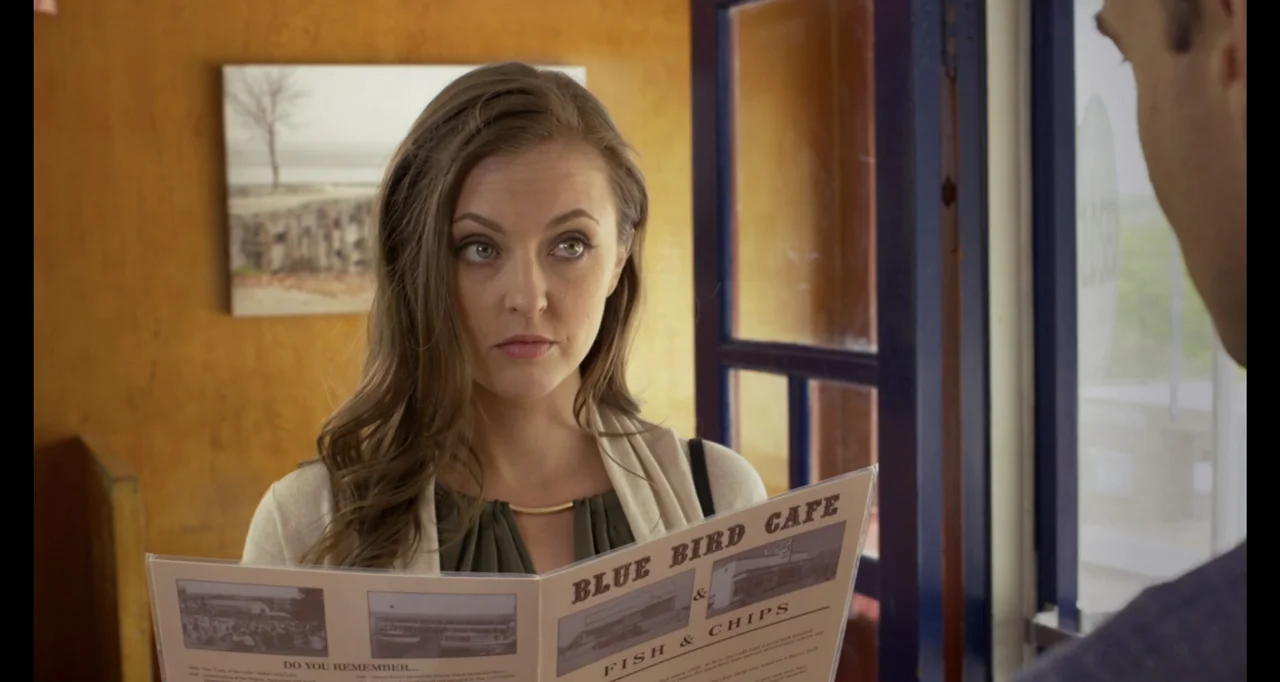 A.R.C.H.I.E. (2016) movie still: medium — An interior cafe with warm wood-paneled walls and a framed print. A woman holds an open menu, speaking to…