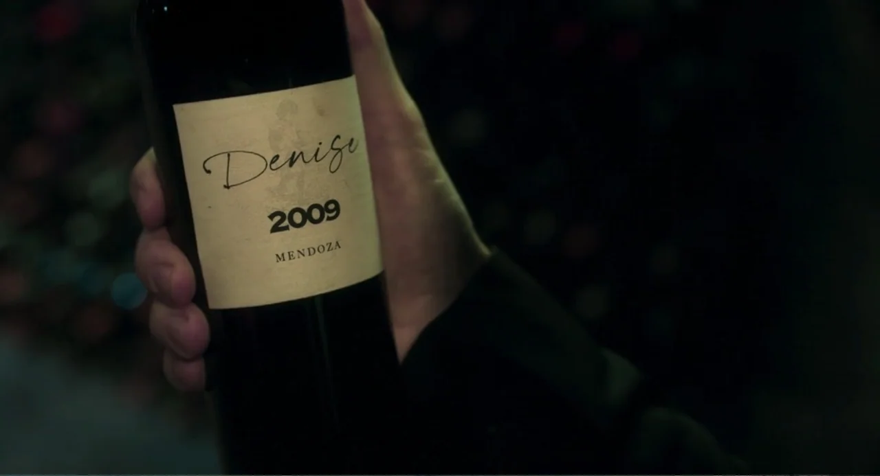 Message in a Bottle (1999) movie still: closeup — A wine bottle with a cream-colored label sits in a dimly lit room. The hand of a person holds the…