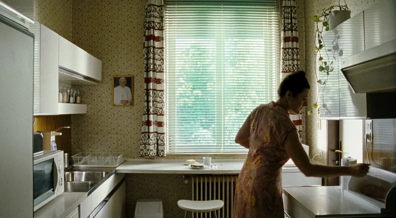 Paradise: Faith (2012) movie still: medium — A modest kitchen features floral wallpaper and a window covered by horizontal plastic blinds. A woman…