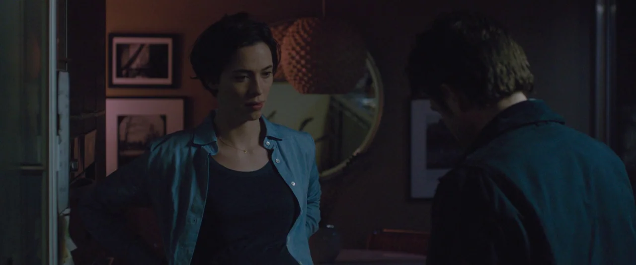The Gift (2015) movie still: medium — A dimly lit room features framed prints on the wall and a circular mirror. A woman faces a man standing with…