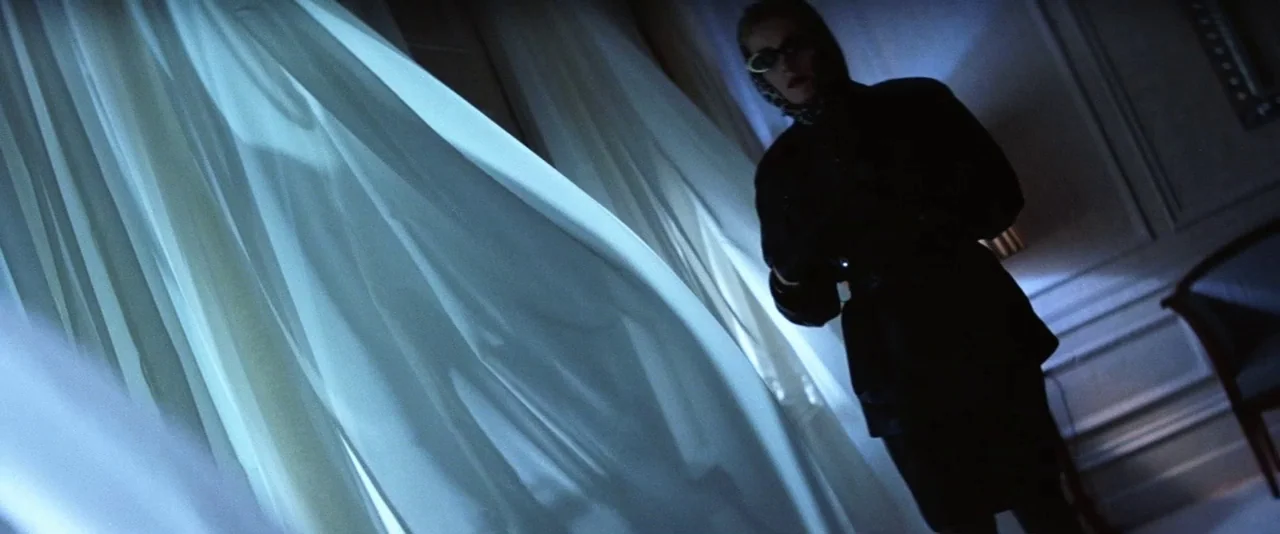 The Hunger (1983) movie still: medium — A dimly lit hotel room features floor-to-ceiling windows covered by billowing white sheer curtains. A woman…