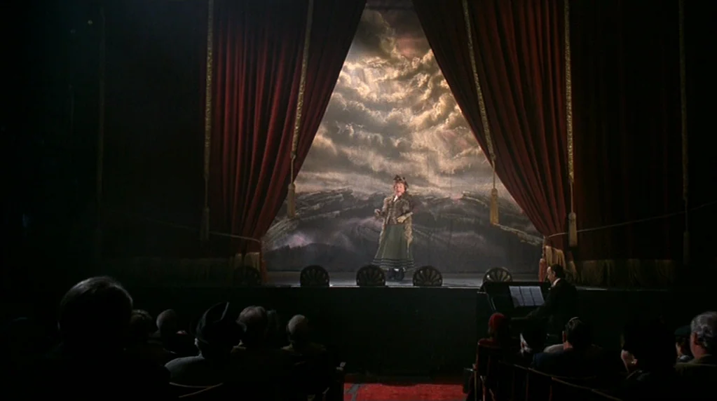 Velvet Goldmine (1998) movie still: wide — An ornate theater stage features a large painted backdrop of dramatic stormy clouds. A single performer…