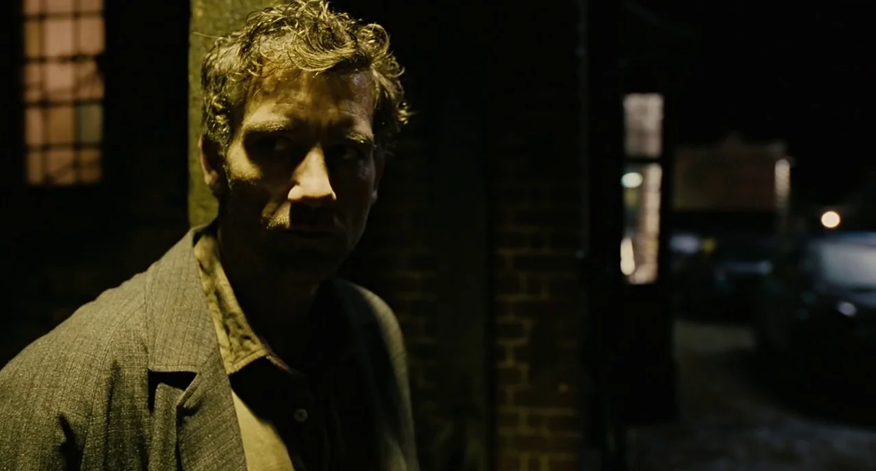 Children Of Men (2006) movie still: medium — A man stands outdoors at night beside the corner of a weathered brick building. He looks away from the…