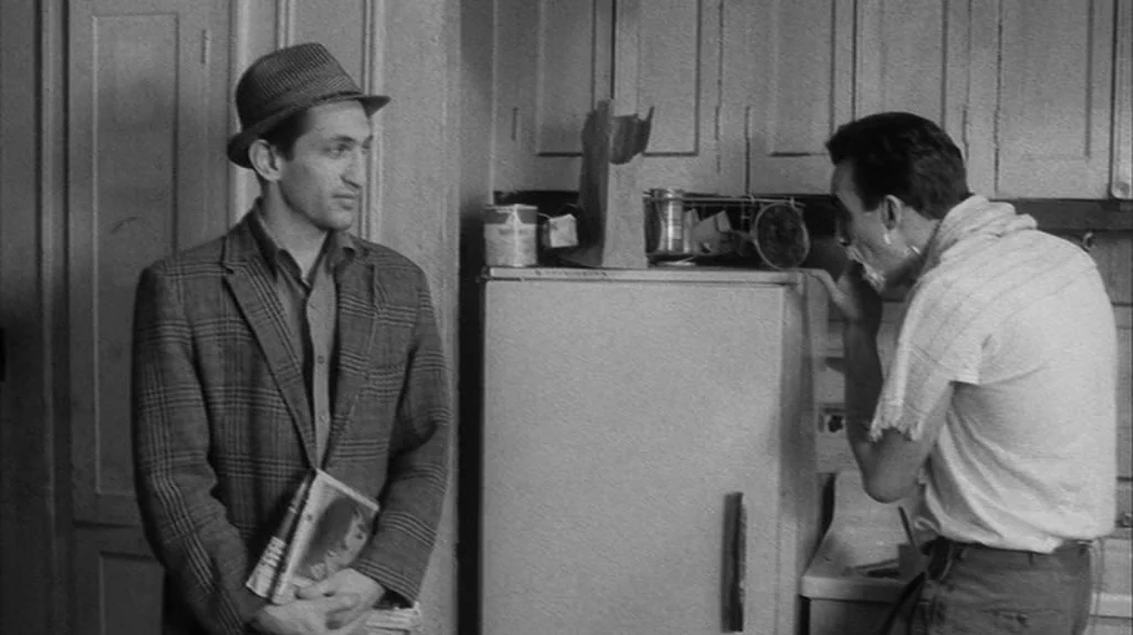Stranger Than Paradise (1984) movie still: medium — A modest domestic kitchen with wooden cabinetry and a classic retro refrigerator. One man stands…