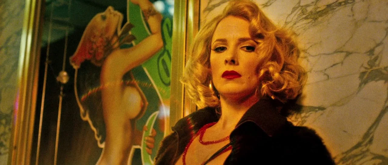 Filth (2013) movie still: medium — A dark, dimly lit venue with a white marble wall and gold-framed signage. A blonde woman with styled hair and red…