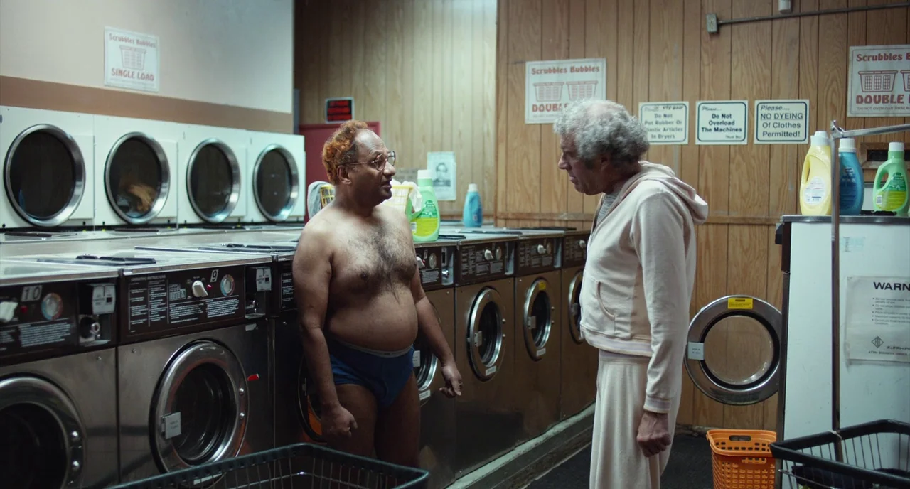 An Evening With Beverly Luff Linn (2018) movie still: medium — A dated laundromat features rows of industrial stainless steel machines against…