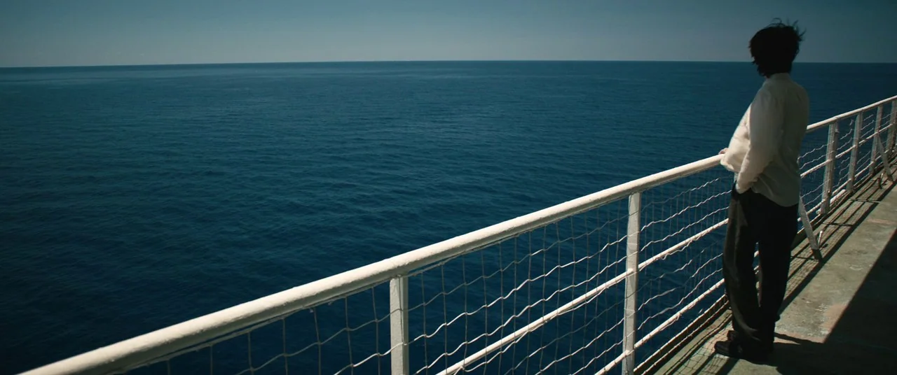 The Rhythm Section (2020) movie still: wide — A metal railing separates a person from the vast expanse of the open ocean. A man stands on the deck…