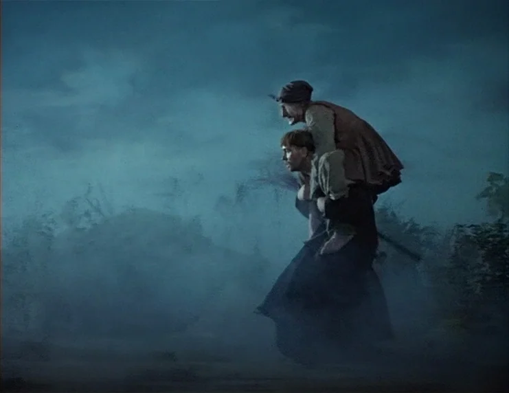 Viy (1967) movie still: fullbody — A dark, misty, open field is illuminated in a deep blue twilight hue. A man carries a smaller elderly person on…
