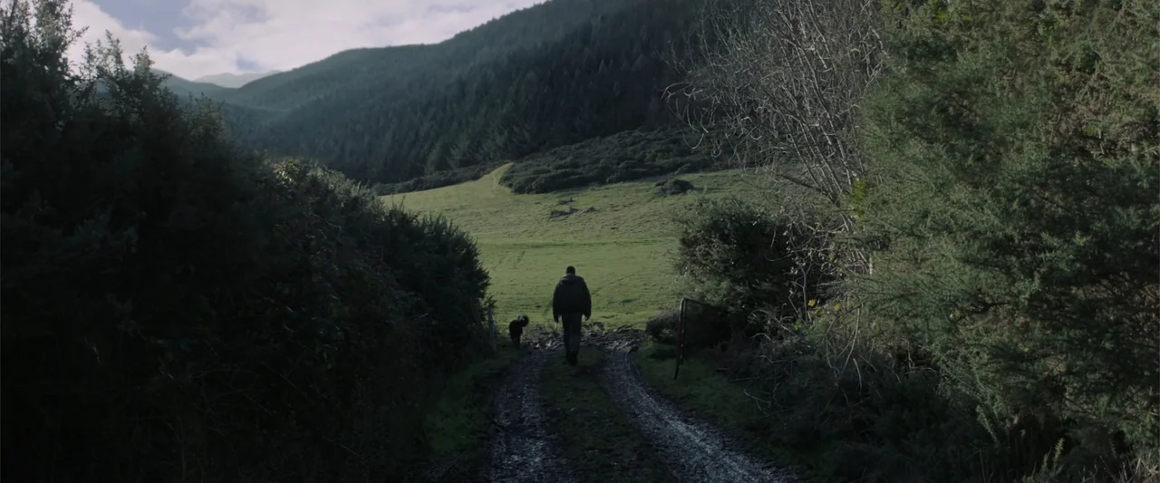 Bring Them Down (2025) movie still: wide — A muddy track leads through dense green foliage toward a rolling hill. A man and a small dog walk away…