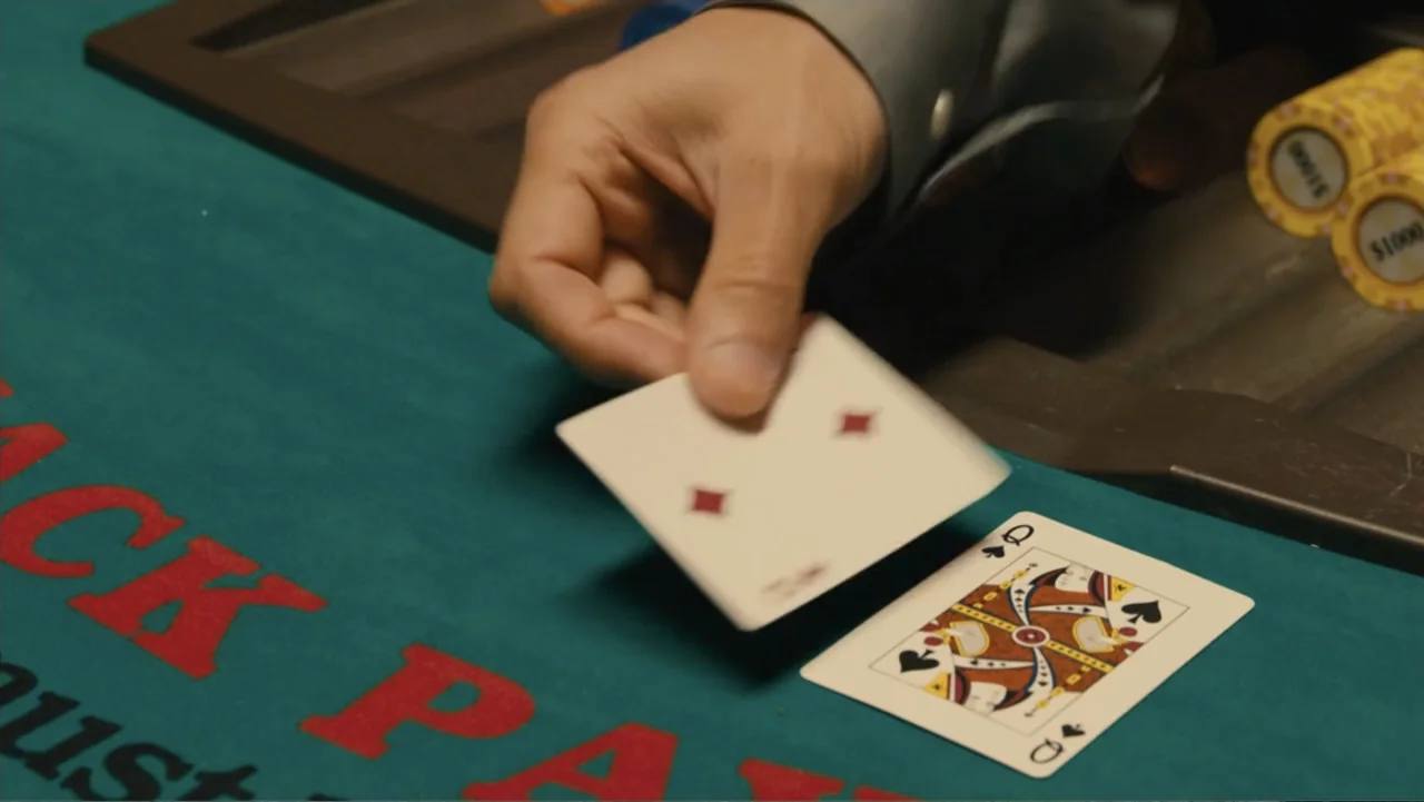 Bleed for This (2016) movie still: closeup — A green felt blackjack table features a hand placing a playing card down. The hand rests near a queen…