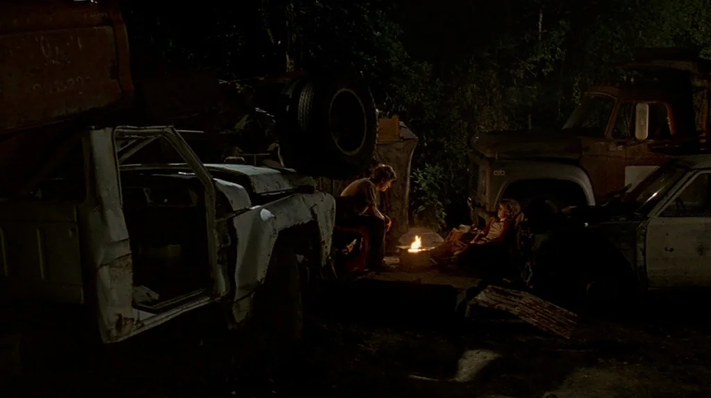 Undertow (2004) movie still: wide — A dark junkyard is filled with rusted, salvaged truck parts and abandoned vehicle frames. Two people sit quietly…
