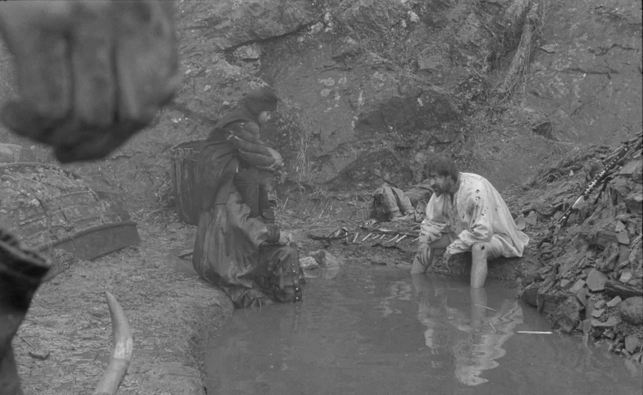 Hard To Be A God (2013) movie still: wide — A rocky, barren landscape contains a small, stagnant pool of muddy water. Two men sit inside the shallow…