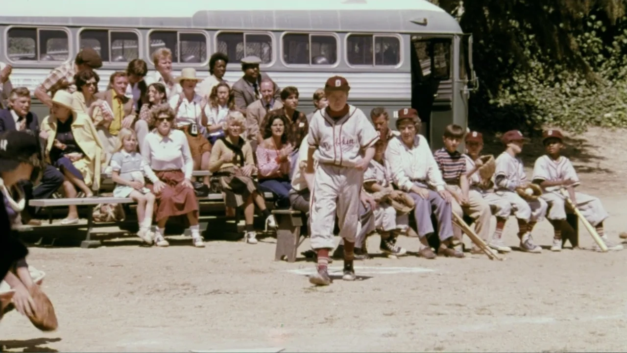 Aunt Mary (1979) movie still: fullbody — A sunny baseball field is filled with spectators sitting on wooden bleachers near a bus. A young boy in a…