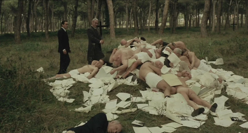 Todo Modo (1976) movie still: wide — A group of older men lie on the ground in a pine forest, partially covered by scattered sheets of paper. Two…